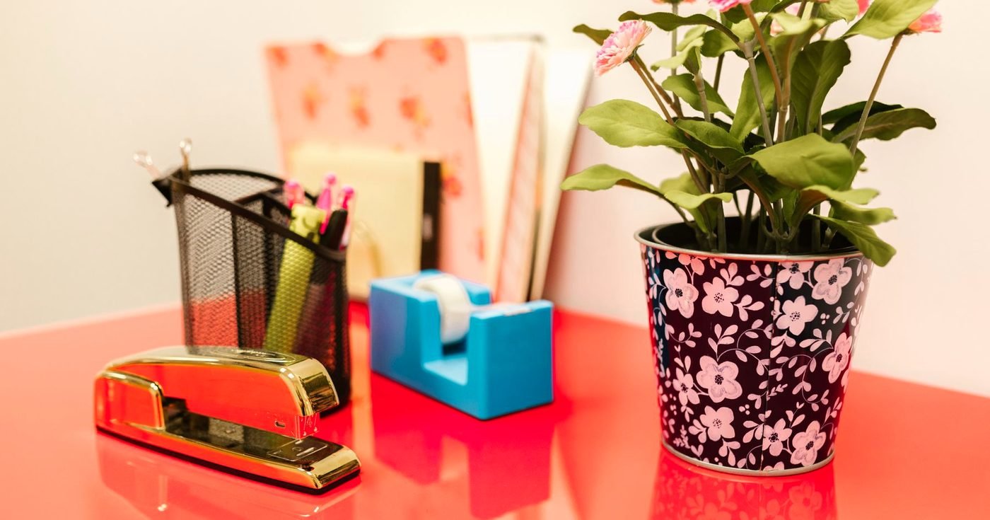 Escritorio rojo con una engrapadora dorada, un portalápices, una cinta adhesiva azul y una maceta con flores