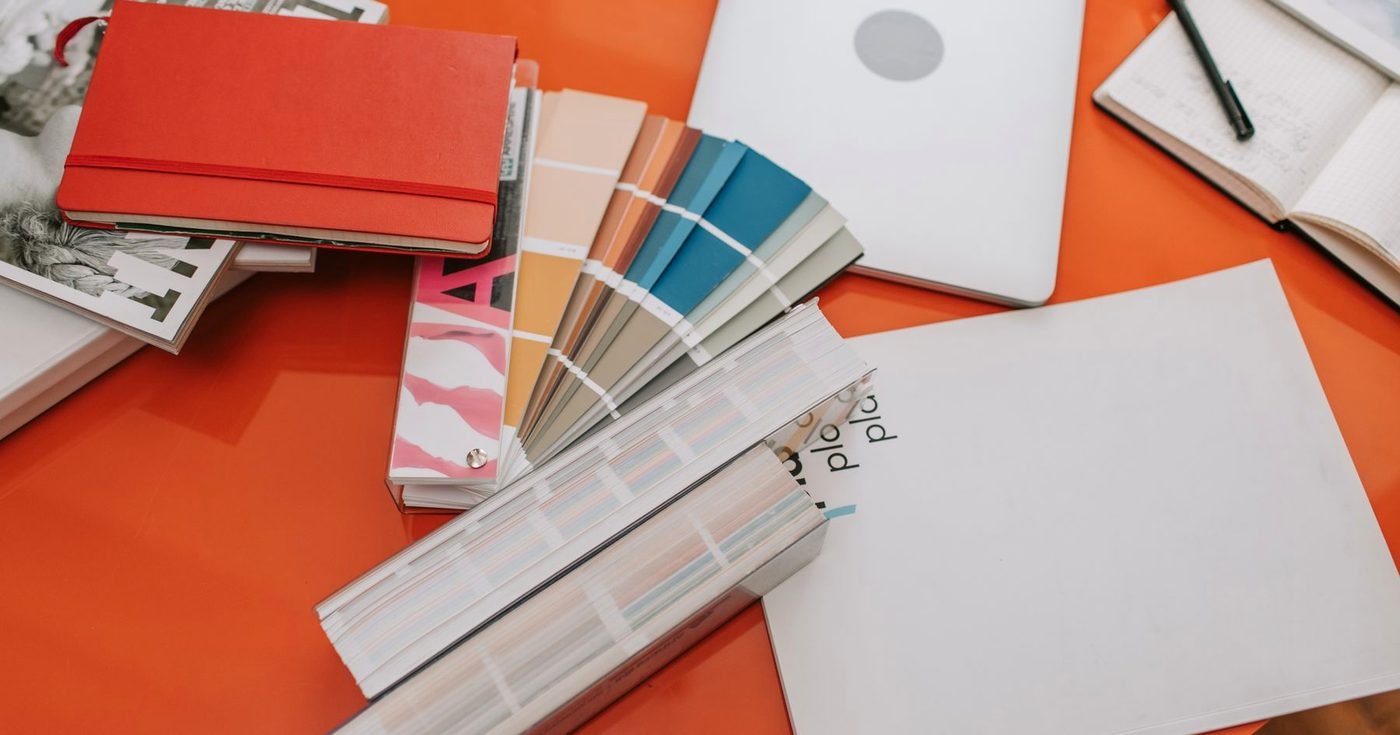Muestrario de colores, cuaderno rojo y hojas sobre un escritorio naranja